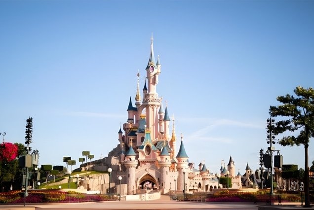 Pink and blue Sleeping Beauty Castle at Disneyland Paris under a clear sky with manicured gardens in the foreground.