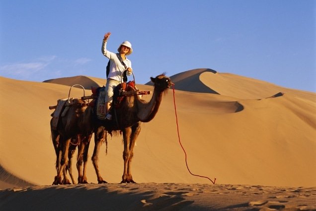 A tourist waving while riding a camel across golden sand dunes during a UAE desert safari tour.