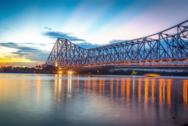 Iconic Howrah Bridge cantilever structure over the Hooghly River in Kolkata under a soft evening sky with city lights.
